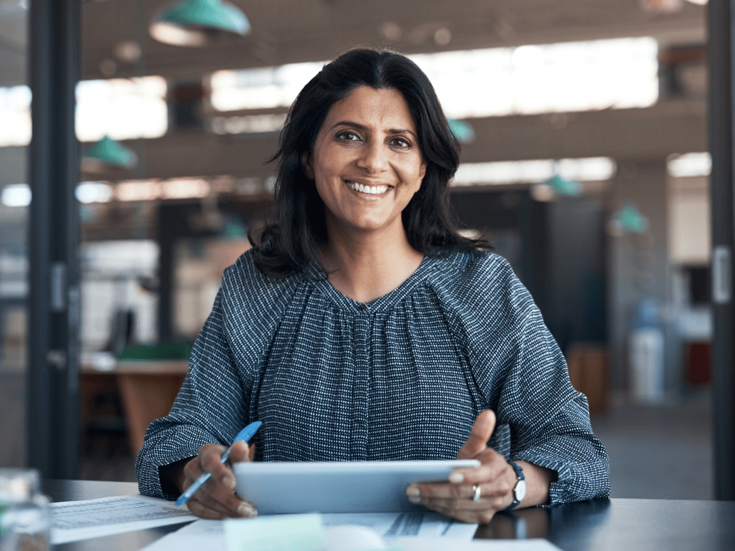 A woman is sitting at a desk holding a tablet and smiling