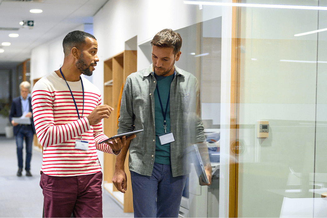 Two men walk down a hallway together, talking and looking at a tablet